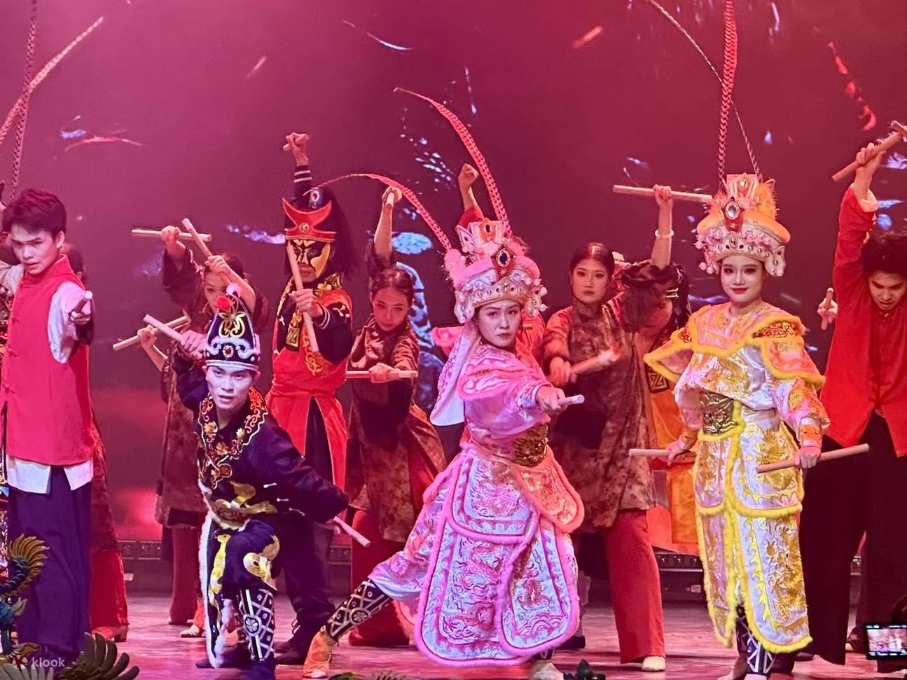 Les acteurs, vêtus de costumes d'opéra et tenant des bâtons, accompagnent la pièce. Les costumes d'opéra jaune pâle sont associés à un éclairage rouge, et les mouvements nets mettent en valeur la tension dramatique de la performance.